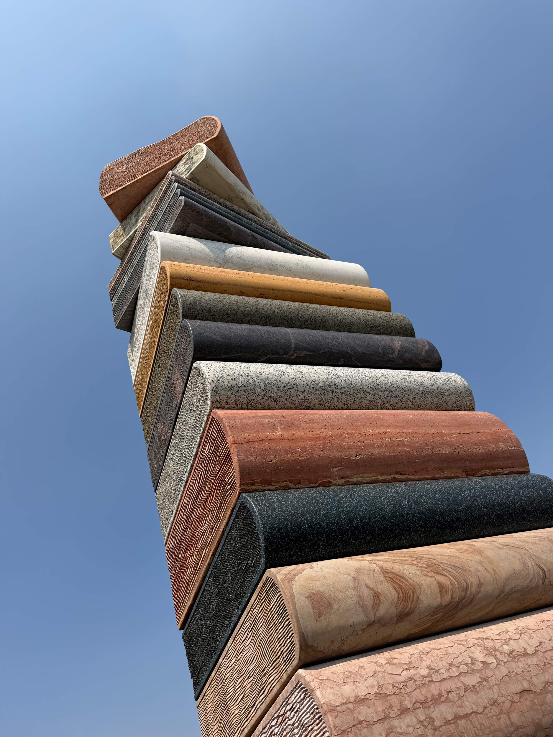 Turm aus übereinander gestapelten, buchähnlichen Steinskulpturen in verschiedenen Gesteinsarten und Farben, von unten fotografiert vor blauem Himmel.