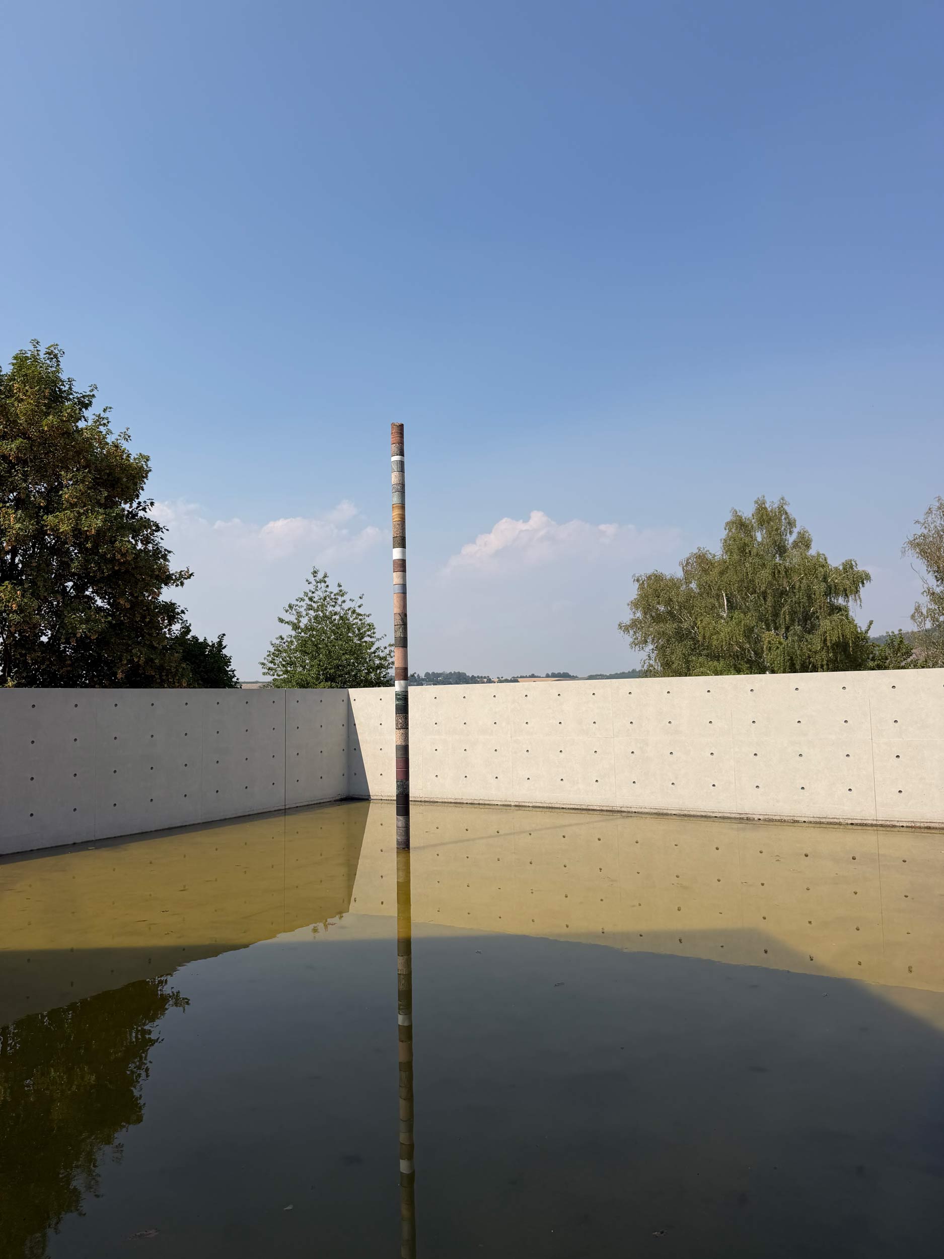 Schlanke, gestreifte Steinsäule steht mittig in einem flachen Wasserbecken vor einer Sichtbetonmauer und spiegelt sich im Wasser.