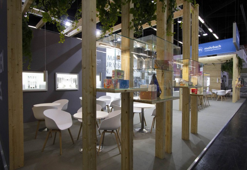 Messestand für Mohrbach auf der Fachpack, Nürnberg 2019, Glasvitrinen, Holzstruktur, Lounge, Bar, Leuchtlogos, Diakästen